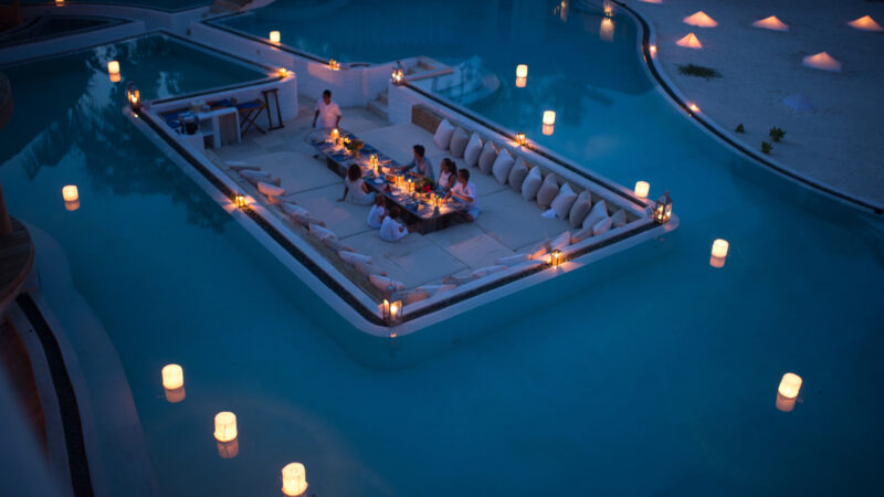 A group of people are seated at a dinner table surrounded by water in a swimming pool at night at the Maldives Luxury Resort. The area is illuminated with numerous lanterns, creating a serene and intimate ambiance. They appear to be enjoying a meal together in this unique, floating setup.