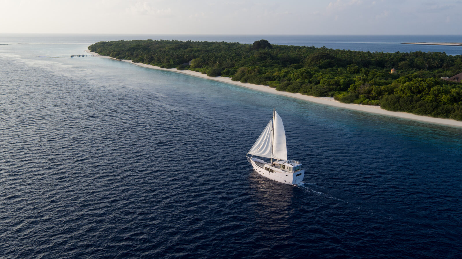 A luxury yacht with white sails gliding over deep blue ocean water near a lush, green island with a sandy shoreline under a cloudy sky, embodying the serene essence of a Maldives holiday.