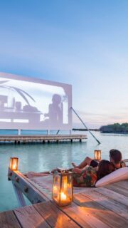 A couple relaxes on a wooden deck over turquoise water, watching a movie projected on a large screen at sunset. Illuminated with lanterns, the cozy, romantic atmosphere near the serene tropical shoreline is part of Soneva's exclusive offers.