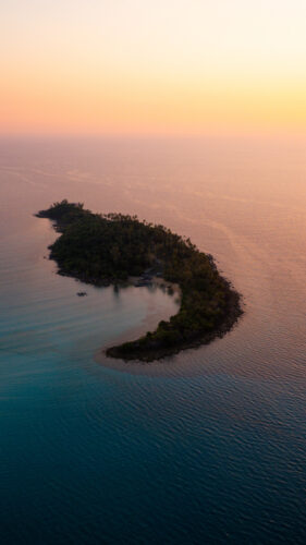 Aerial view of a small, elongated island surrounded by calm, turquoise waters during sunrise or sunset. The sun is low on the horizon, casting a golden reflection on the sea. This Thailand exclusive experience showcases an island covered with dense greenery.