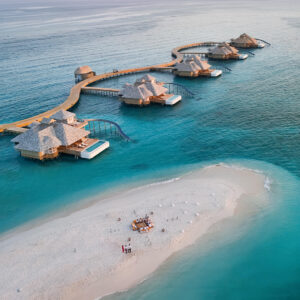 Aerial view of luxurious overwater bungalows connected by wooden walkways extending into the turquoise ocean at a Maldives private resort. In the foreground, a small sandy beach hosts a group with a picnic setup, surrounded by sun umbrellas. The serene sky is subtly lit with warm hues.
