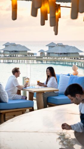 A couple dines at a seaside restaurant with wooden decor and blue cushions, enjoying drinks while a waiter prepares a dish at their table. Overwater bungalows of the Maldives luxury resort are illuminated by the sunset over the ocean, creating a perfect backdrop for this romantic evening.
