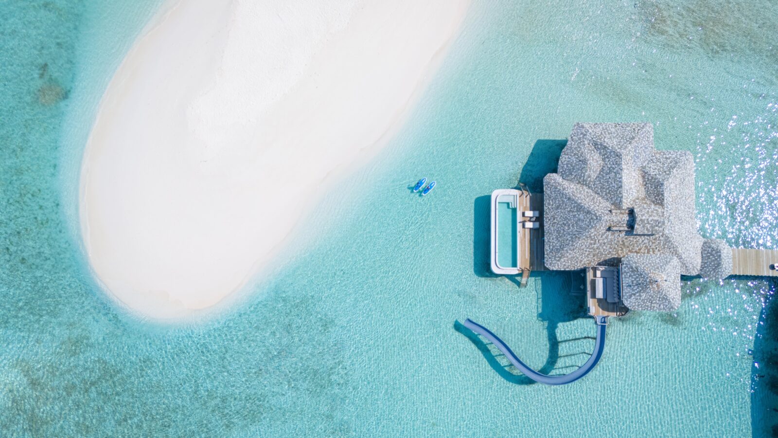Aerial view of a tropical overwater bungalow at a Maldives luxury private resort, featuring a slide leading into clear turquoise waters. A small white sandbar is visible nearby. Two people in blue swimwear are floating in the water beside the bungalow. The scene is serene and picturesque.