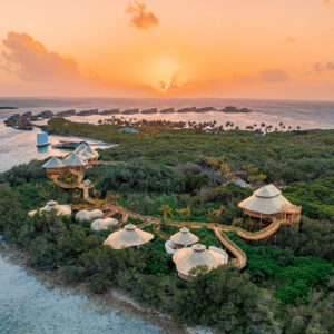 A scenic aerial view of an island resort during sunset. The resort, featuring several thatched-roof huts amidst lush greenery and connected by wooden walkways, glows under the sun's golden hue. Discover Soneva Exclusive Offers as the sun sets over the ocean, casting its warm light on the tranquil waters.