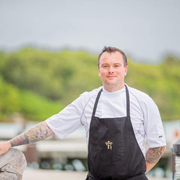 A chef with tattoos, wearing a white shirt and black apron, stands outdoors with one hand on a large piece of driftwood. The background features greenery and a soft-focus sky.