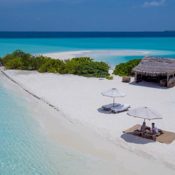 A small, secluded tropical island with a thatched-roof hut and vibrant green shrubs. Two people sit under white umbrellas on the pristine white sand next to the clear turquoise ocean. The bright blue sky enhances this tranquil, picturesque Soneva experience in the Maldives.
