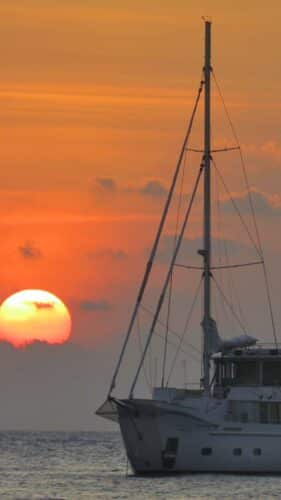 A luxurious white sailboat is anchored on the calm waters of the Maldives during a vibrant sunset. The sun, partially obscured by light clouds, casts an orange and pink glow across the sky and reflects on the water's surface, epitomizing a luxury yacht holiday at Soneva.