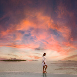 A person in a white outfit walks along a pool's edge with their reflection visible in the water. Behind them, the vast ocean and a small island form a picturesque backdrop against a dramatic sunset sky. Experience moments like this with Soneva Exclusive Offers, where luxury meets tranquility.