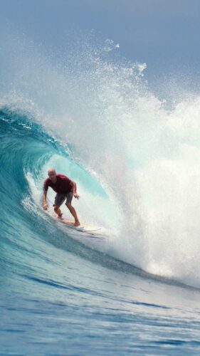 A surfer in a red shirt rides the inside of a large, blue wave as it curls overhead, with white foam spraying around. The ocean is clear and the sky is slightly visible in the background. The surfer maintains a low stance, capturing the thrill of the moment during their Maldives Luxury Yacht Holiday at Soneva.