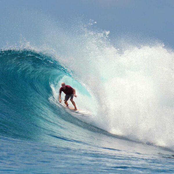 A surfer in a red shirt rides the inside of a large, blue wave as it curls overhead, with white foam spraying around. The ocean is clear and the sky is slightly visible in the background. The surfer maintains a low stance, capturing the thrill of the moment during their Maldives Luxury Yacht Holiday at Soneva.