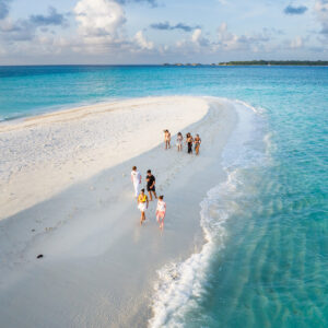 A group of people walks along a narrow strip of white sandy beach surrounded by turquoise water under a partly cloudy sky. Some are taking photos, while others leisurely stroll, perhaps even reflecting on Soneva Exclusive Offers. The beach tapers off into the ocean, creating a picturesque scene.