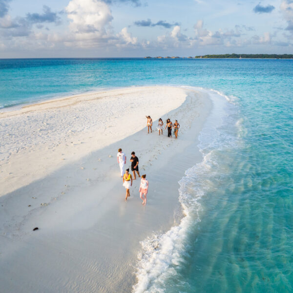 A group of people walks along a narrow strip of white sandy beach surrounded by turquoise water under a partly cloudy sky. Some are taking photos, while others leisurely stroll, perhaps even reflecting on Soneva Exclusive Offers. The beach tapers off into the ocean, creating a picturesque scene.