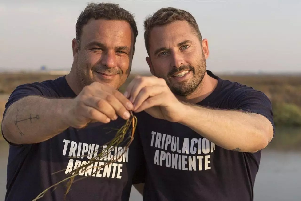 Two smiling individuals in navy shirts with "TRIPULACIÓN APONIENTE" printed on them, standing outdoors. They hold a small plant together, with a natural landscape in the background.