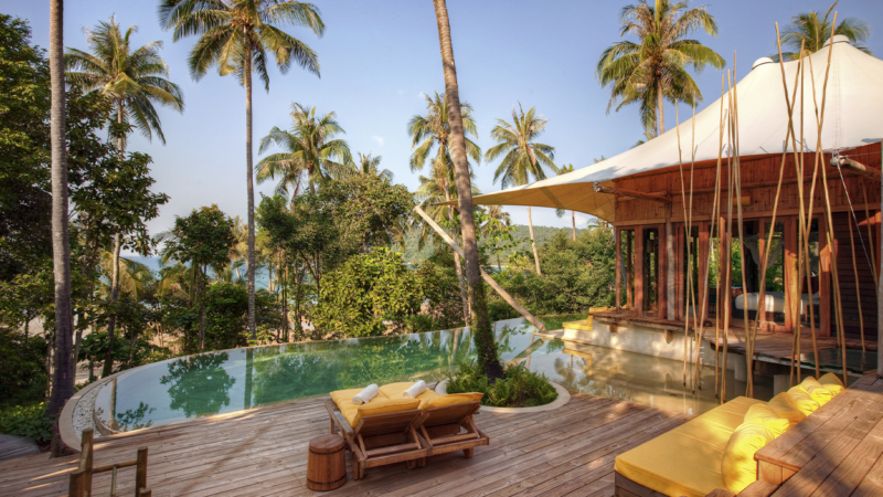 A serene tropical retreat at Soneva Kiri Resort features a private pool surrounded by lush greenery and palm trees. A wooden deck with yellow cushioned chairs overlooks the pool. Nearby, a tented pavilion with open sides enhances the tranquil, luxurious ambiance.