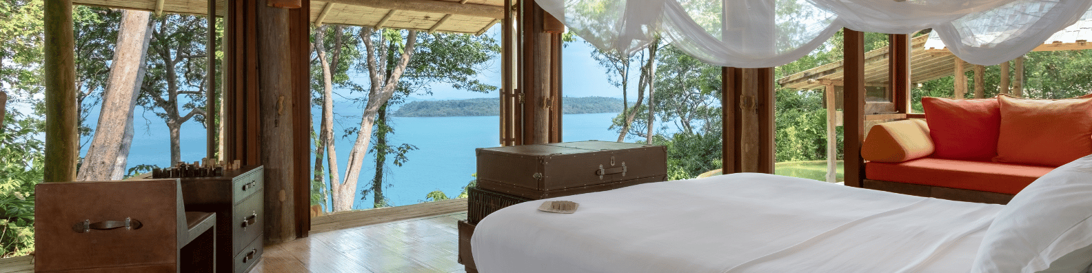 A cozy bedroom with a canopy bed beside large windows offering a stunning view of the ocean and surrounding forest. The room features wooden furniture, including a chest and a nightstand. The bed is made with white linens, and a red sofa is in the corner—truly a Thailand exclusive experience at Soneva.