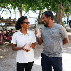 Two people stand outdoors under trees in the picturesque Maldives. They smile at each other while holding glass bottles. The person on the left wears a white shirt and dark sunglasses, and the person on the right wears a gray shirt and dark pants. Bicycles and motorbikes are in the background, adding to the Soneva Namoona vibe.