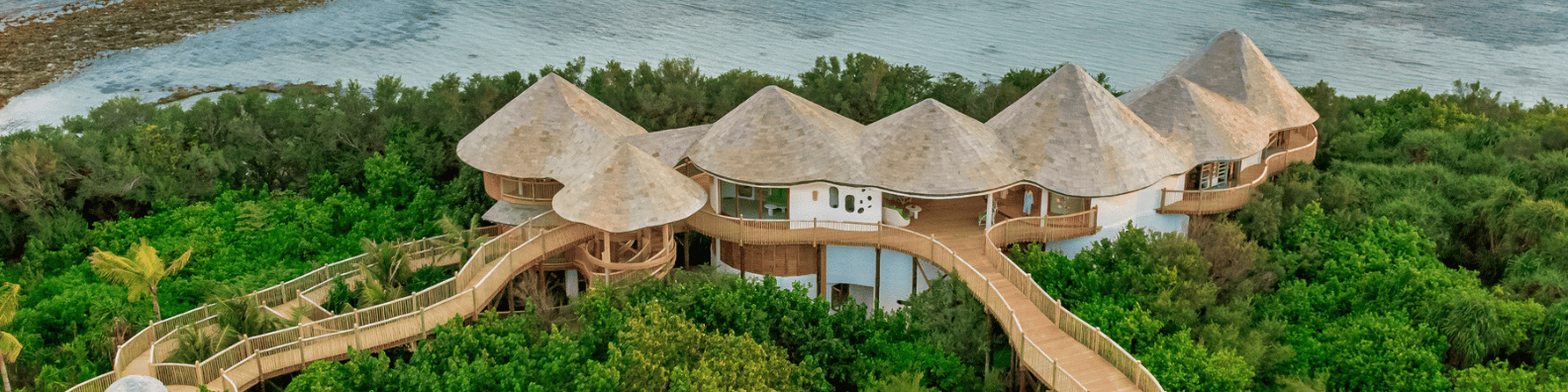 Aerial view of an expansive, circular villa with multi-level thatched roofs nestled among dense greenery on a coastal landscape. Elevated wooden walkways connect various parts of the Maldives wellness retreat, and the ocean is visible in the background.