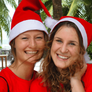 Two smiling women wearing red Santa hats and red shirts stand close together, with tropical palm trees and a beach in the background. They seem to be enjoying a festive Family Holiday in the Maldives, savoring a joyful moment at Soneva&#039;s luxurious resort.