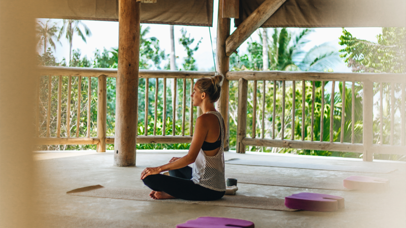 A person sits cross-legged in a meditative pose on a yoga mat inside an open wooden pavilion with a view of lush greenery and palm trees, reminiscent of a serene Maldives spa. Cushions and yoga props are scattered around them on the floor, enhancing the overall wellness experience.