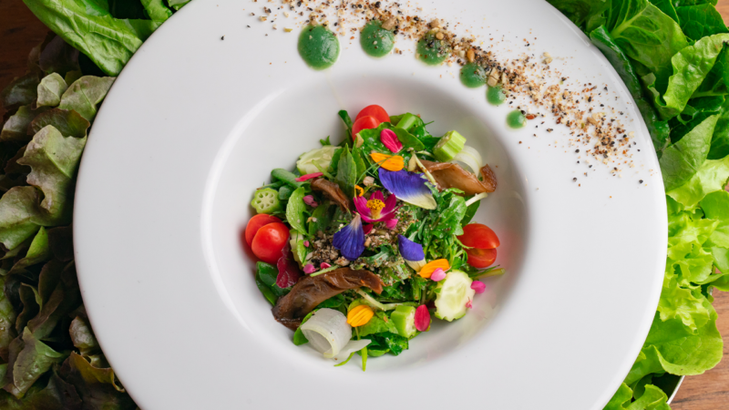 A vibrant salad served on a white plate at Soneva Kiri Resort. The salad includes cherry tomatoes, green leaves, edible flowers, and various vegetables. The plate is garnished with dots of green sauce and a sprinkle of seasoning. Fresh greens surround the plate.
