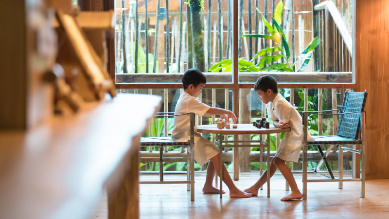 Two children wearing white outfits sit at a round table by a large window, engaged in a playful activity. The room has wooden walls and a cozy atmosphere, reminiscent of a family holiday. With lush greenery visible outside the window, it feels as inviting as Soneva in the Maldives.