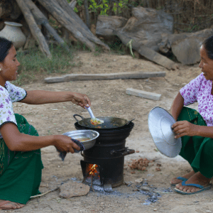 Two women are sitting outdoors, cooking together over a small fire. One woman stirs food in a pot on a metal stove, while the other holds a lid. Both are dressed in green skirts and floral tops, surrounded by a rustic setting with wood and greenery, reminiscent of Maldives Namoona traditions.