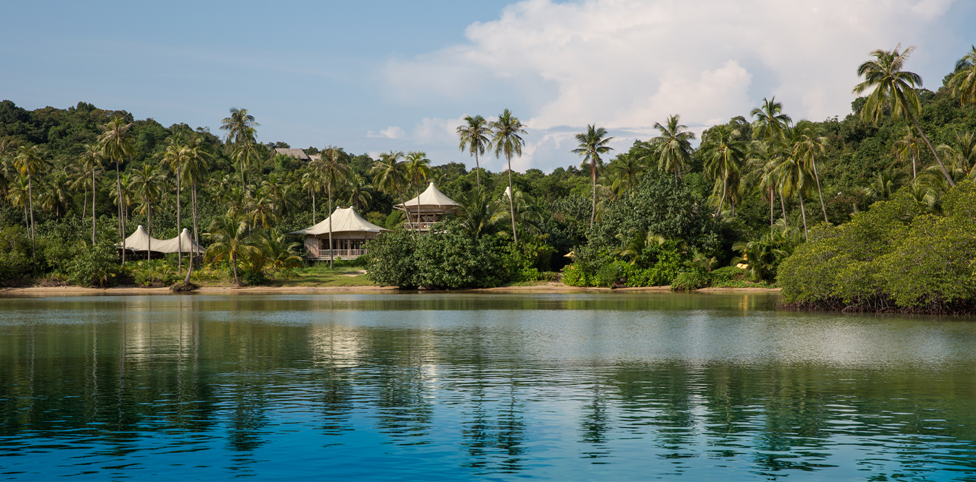 A serene tropical scene featuring a calm body of water reflecting the lush greenery and palm trees on the shore. In the background, a few thatched-roof buildings, characteristic of Soneva Kiri Resort, are nestled among the trees under a partly cloudy sky.
