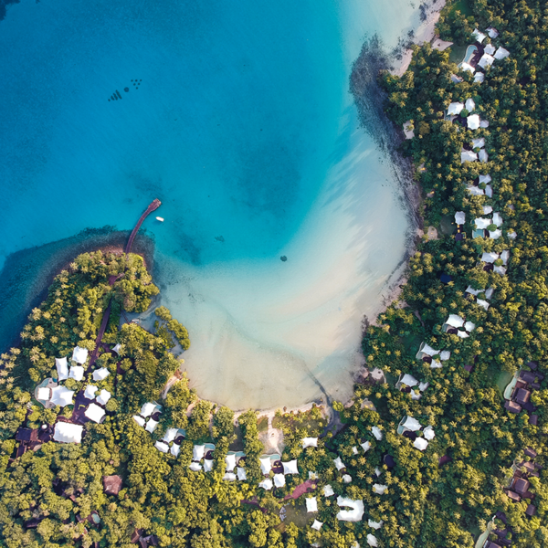 Aerial view of a stunning tropical beach with turquoise waters gently meeting the sandy shore. Lush greenery surrounds the beach, with white-roofed buildings scattered among the trees. A small pier extends into the water from one side of the coastline, reminiscent of Soneva Kiri Resort's charm.