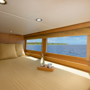 A cozy bed with beige linens inside a luxury yacht features a tray with a glass bottle and cup. Large windows offer a scenic view of the Maldives' ocean and a distant tropical island under a clear blue sky. Recessed lighting illuminates the interior, enhancing your holiday experience.
