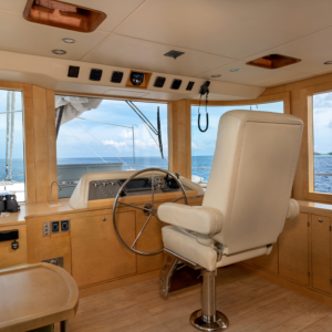 The interior helm station of a luxury yacht, perfect for a Maldives holiday, features a large white captain's chair, a wooden steering wheel, and various control panels. The view out the wide windows showcases a clear, sunny day with a calm ocean and blue sky in the background.