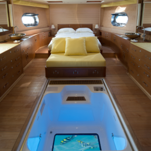 The interior of a luxurious yacht's cabin features a bed with yellow bedding and pillows, wooden cabinetry, and two circular windows. There's a glass floor panel revealing an aquarium beneath, showcasing fish swimming in the illuminated water below—a perfect setting for your Maldives Luxury Yacht Holiday.