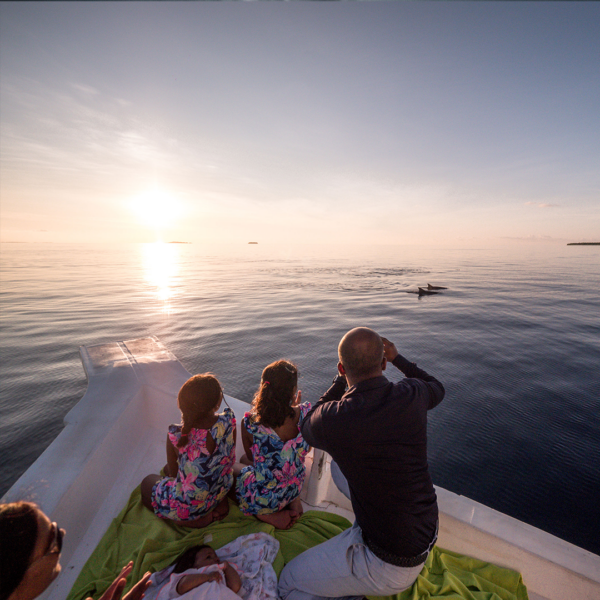 A family is on a boat during sunset, facing the horizon. Two children in matching floral dresses and an adult watch dolphins in the water, enjoying Soneva Exclusive Offers. An infant rests on a blanket nearby. The sky is clear and the water is calm.