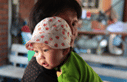 A baby wearing a white hat with red polka dots and a green shirt is being carried by an adult. In the blurred background, reminiscent of a family holiday in Soneva, rustic indoor items and utensils are scattered about, evoking memories of simpler times.