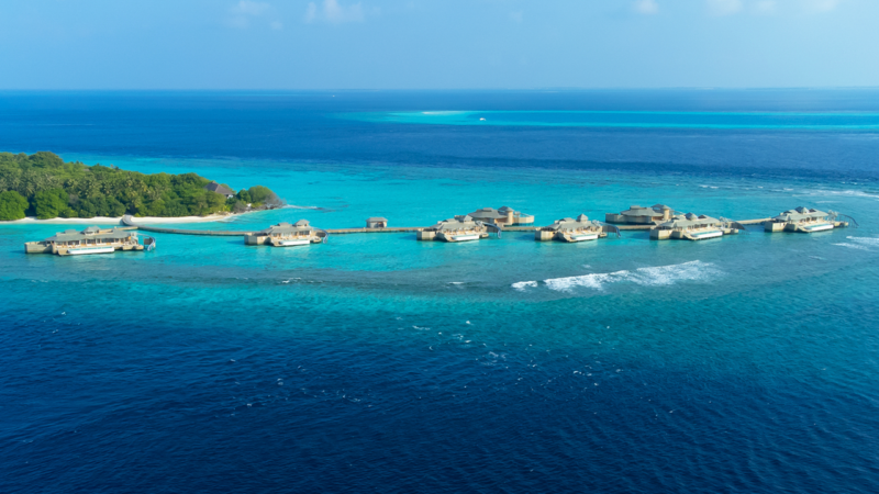 Soneva Fushi 别墅鸟瞰图，这是马尔代夫一个小岛上的豪华度假村，建在碧蓝的海水之上，四周环绕着深蓝色的海洋。度假村建筑之间似乎有人行道相连，在明净的天空下，部分岛屿绿意盎然。