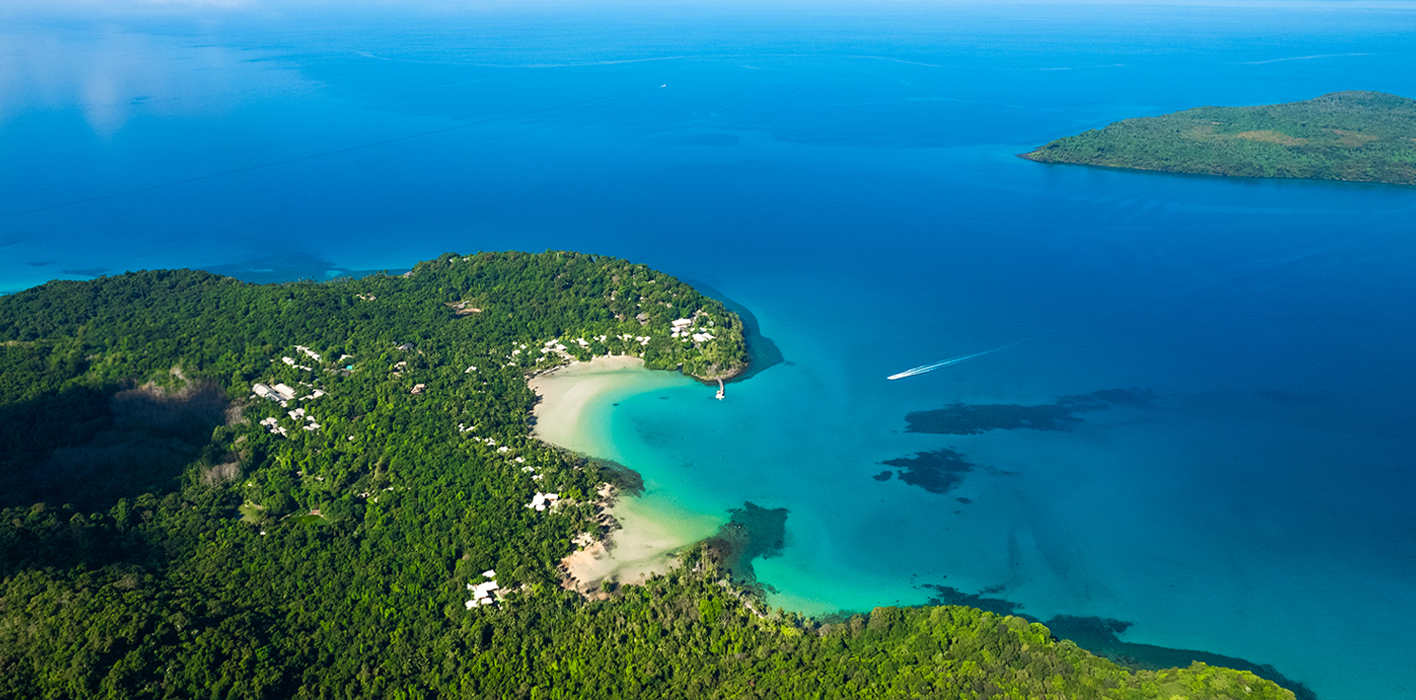 Aerial view of a tropical island near Soneva Kiri Resort with lush greenery, white sand beaches, and turquoise water. A boat creates a white trail in the sea, and another island is visible in the distance under a clear blue sky.