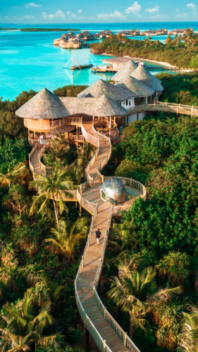 Aerial view of a tropical resort featuring wooden pathways weaving through lush green foliage, leading to elevated round villas overlooking a turquoise ocean. In the distance, additional villas on stilts over the water. A person walks along the pathway, embodying the Maldives exclusive experience at Soneva.