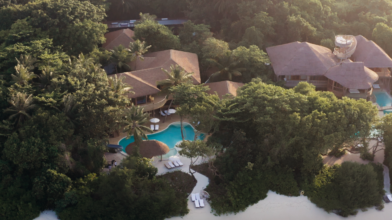 Aerial view of Soneva Residences, a tropical resort surrounded by dense greenery. Several thatched-roof buildings are nestled among the trees, along with a pool area featuring lounge chairs and umbrellas. A sandy beach lies adjacent to the resort, leading to clear turquoise waters.