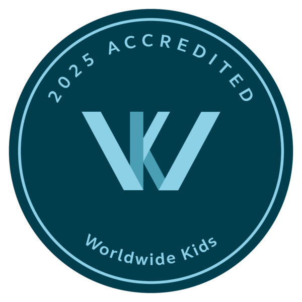 A circular emblem with the text &quot;2025 Accredited&quot; at the top and &quot;Worldwide Kids&quot; at the bottom sits proudly, reminiscent of a family resort in the Maldives. At its center, a stylized &quot;W&quot; and &quot;K&quot; intertwine gracefully in light blue on a dark teal background.